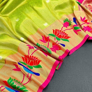 May include: A close-up of a vibrant green saree with intricate floral patterns. The saree features a gold border with red, pink, and blue lotus flower designs. The edges are trimmed with a bright pink border, creating a striking contrast.