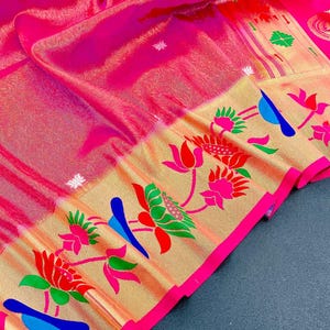 May include: A vibrant pink textile with a gold border featuring floral and bird designs in red, green, and blue. The fabric has a subtle pattern and a bright pink border. This textile is likely a saree or similar garment.