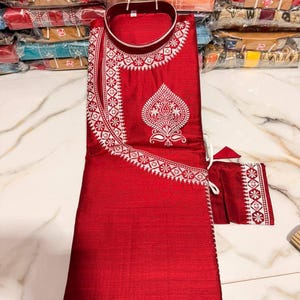Puede incluir: Un conjunto de kurta pijama rojo con intrincados bordados blancos. El kurta presenta un cuello mandarín y patrones detallados a lo largo del escote y la parte delantera. Se incluye una pequeña bolsa a juego. La tela tiene un brillo sutil.