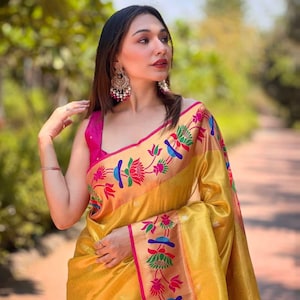 May include: A woman wearing a gold-colored sari with a pink blouse. The sari features a floral pattern in red, blue, and green. The woman is also wearing large, ornate earrings.