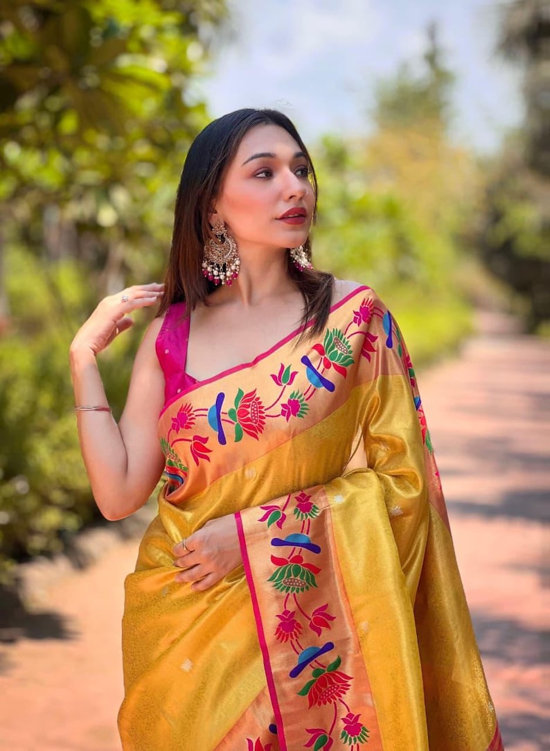 May include: A woman wearing a gold-colored sari with a pink blouse. The sari features a floral pattern in red, blue, and green. The woman is also wearing large, ornate earrings.