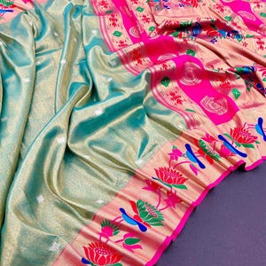 May include: A luxurious, draped saree in shades of teal and gold, with a vibrant pink border. The border features floral and bird motifs in red, green, and blue. The fabric has a subtle sheen, adding to its elegance.