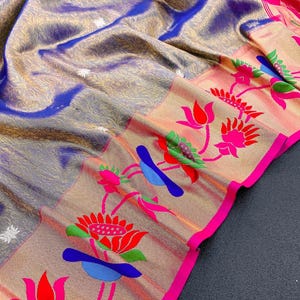 May include: A luxurious textile with a shimmering, textured surface in shades of blue, gold, and gray. The border features a floral pattern with red, pink, blue, and green accents, outlined with a bright pink trim. The fabric is intricately detailed.