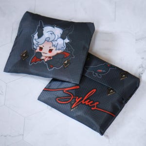 May include: Two black reusable shopping bags with a white and red cartoon character on one bag and a black bird on the other. The bags have a red signature that says "Sylus" on one bag.