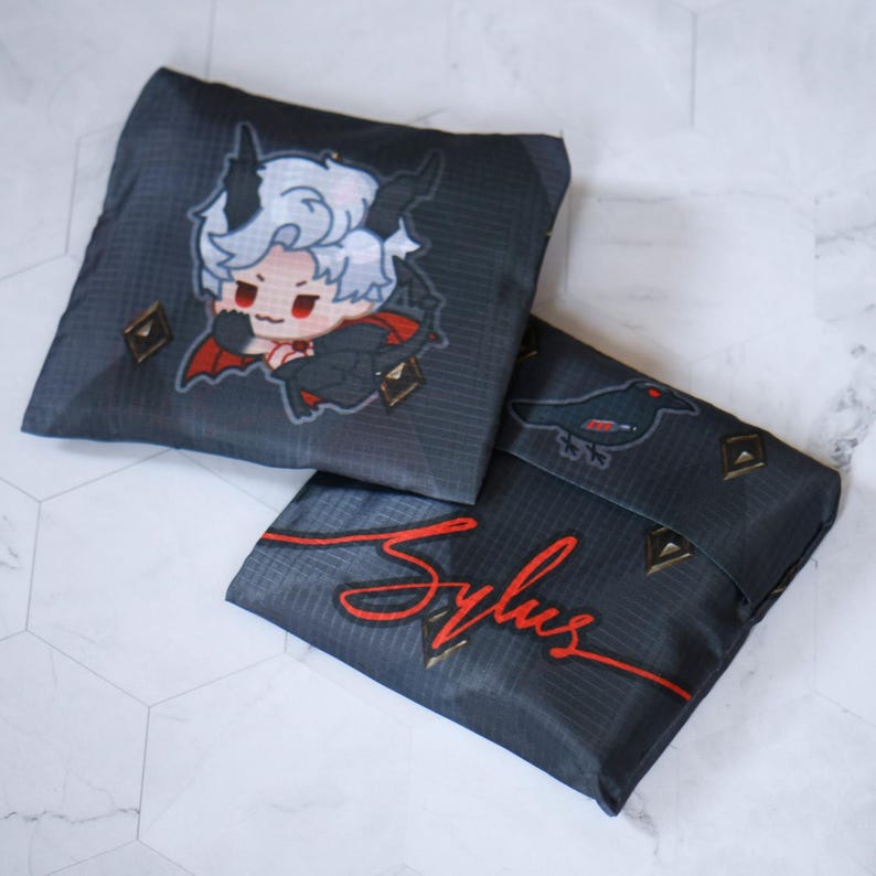 May include: Two black reusable shopping bags with a white and red cartoon character on one bag and a black bird on the other. The bags have a red signature that says "Sylus" on one bag.