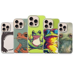 May include: A collection of five phone cases featuring colorful frog illustrations. The designs include a penguin, a frog with a cigarette, a frog giving a peace sign, a frog wearing sunglasses, and a watercolor frog.