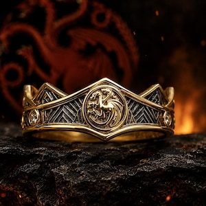 May include: A gold ring with a crown design. The ring features a central circular emblem with a dragon, surrounded by geometric patterns and small, clear gemstones. The ring is set against a dark, textured background.