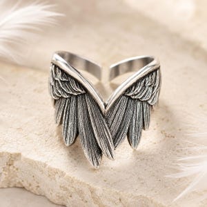 May include: A silver-colored ring with an open design, featuring detailed wing-shaped accents. The wings are intricately textured, with each feather clearly defined. The ring is set against a neutral background, highlighting its metallic sheen.