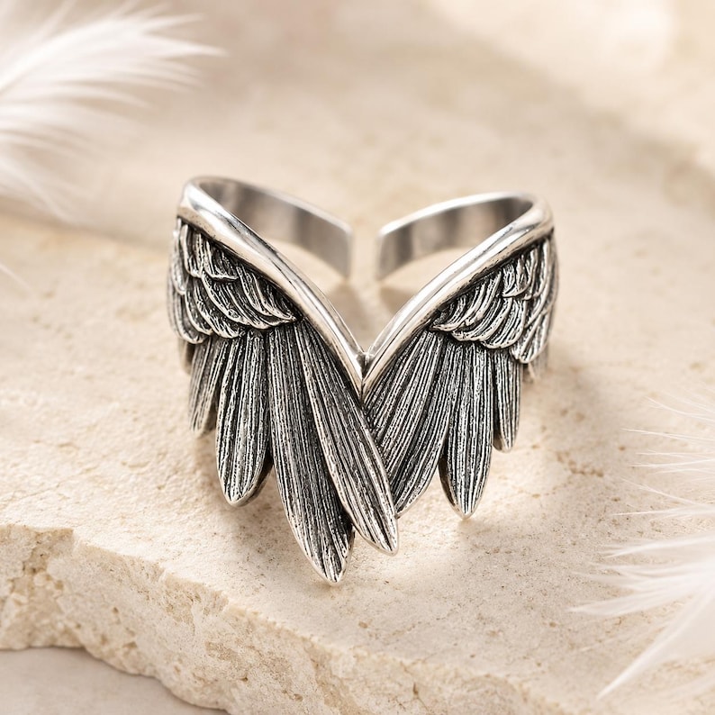 May include: A silver-colored ring with an open design, featuring detailed wing-shaped accents. The wings are intricately textured, with each feather clearly defined. The ring is set against a neutral background, highlighting its metallic sheen.