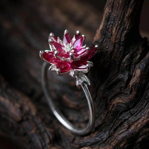 May include: A silver ring featuring a flower design with vibrant red petals and a small, sparkling center stone. The ring's band is slender, and a small leaf detail adds to the floral theme. The ring is set against a dark, textured background.