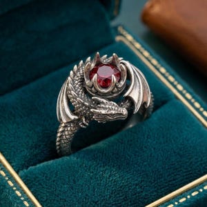 Pode incluir: Um anel de dragão prateado com uma grande pedra preciosa vermelha redonda. O dragão tem escamas, asas e uma cabeça detalhadas. O anel é exibido em uma caixa de anel de veludo verde-azulado.