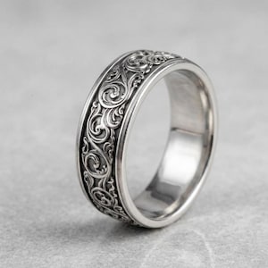 May include: A silver ring with an intricate, swirling floral pattern. The band is wide and the design is embossed around the entire ring. The ring has a polished finish and a classic, elegant style.