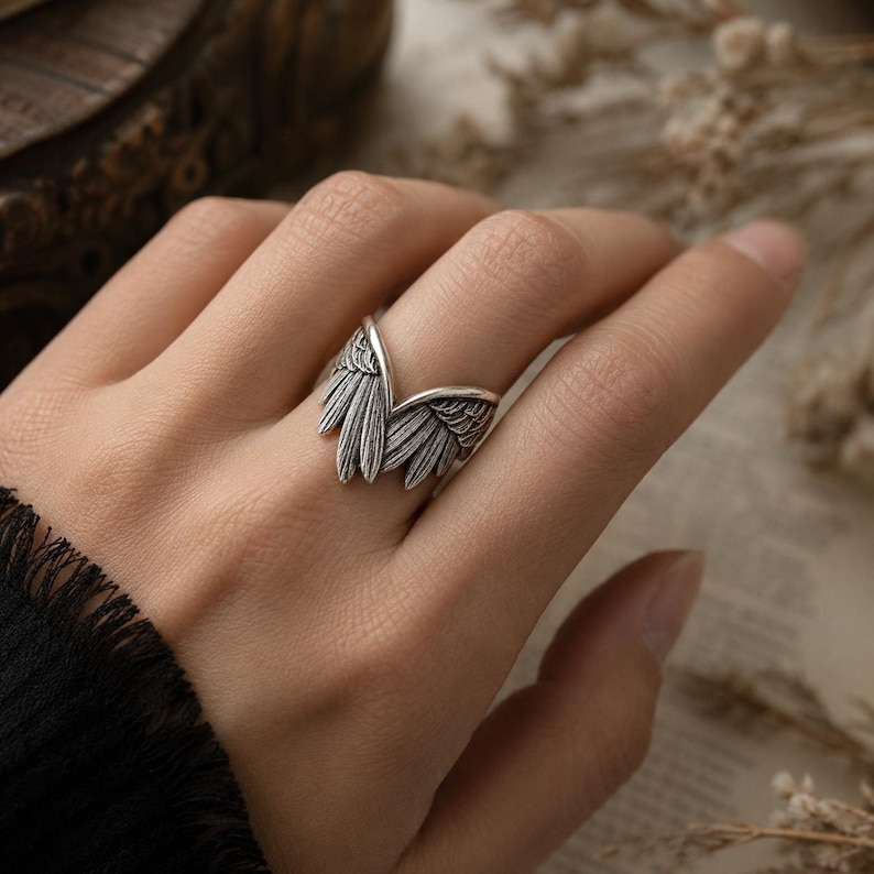 May include: A silver-toned ring featuring a detailed wing design. The ring's wings are spread open, with intricate feather detailing. The ring is worn on a finger, showcasing the jewelry's craftsmanship and design.