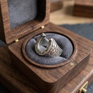 May include: A silver ring shaped like a horseshoe with engraved leaf patterns, resting inside a dark brown wooden box lined with grey velvet. The ring features a prominent U-shape design with detailed metalwork along the outer band surface.