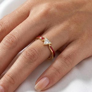 May include: A gold ring with a triangular, clear gemstone and red enamel accents. The ring is on a finger, showcasing its design and craftsmanship. The ring's band features a vibrant red enamel, adding a pop of color to the jewelry.