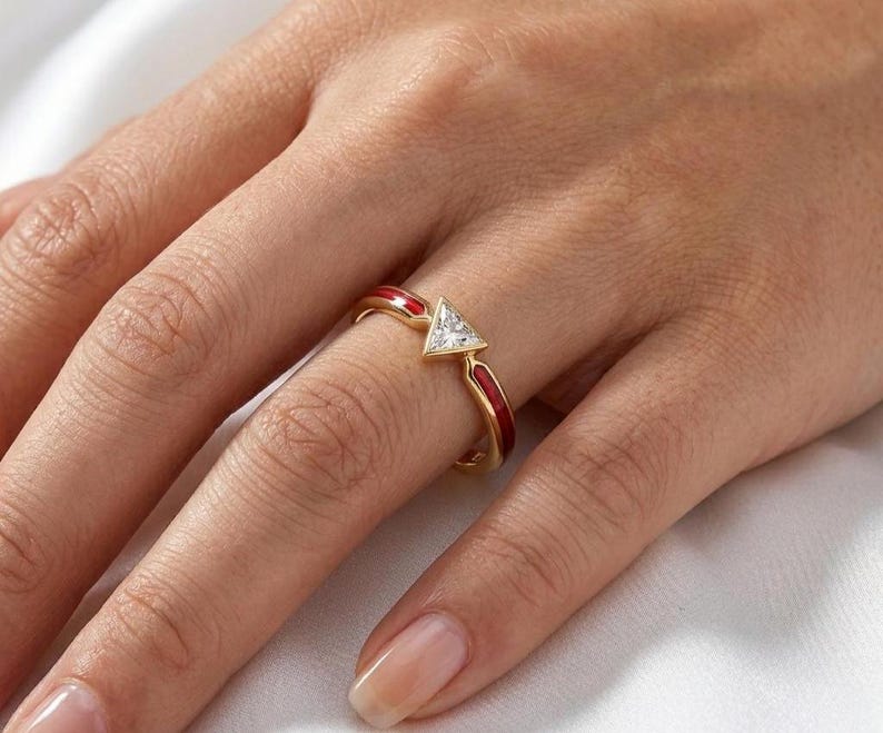 May include: A gold ring with a triangular, clear gemstone and red enamel accents. The ring is on a finger, showcasing its design and craftsmanship. The ring's band features a vibrant red enamel, adding a pop of color to the jewelry.