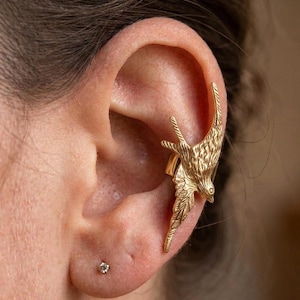 May include: A gold-colored ear cuff shaped like a bird, with detailed feather and wing designs. The cuff wraps around the ear, creating a unique and stylish accessory. A small, clear stud earring is also visible.