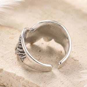May include: A silver-colored adjustable ring with a detailed feather design on one side. The ring has a smooth, polished finish and an open design, allowing for size adjustment. The ring is set against a neutral background.
