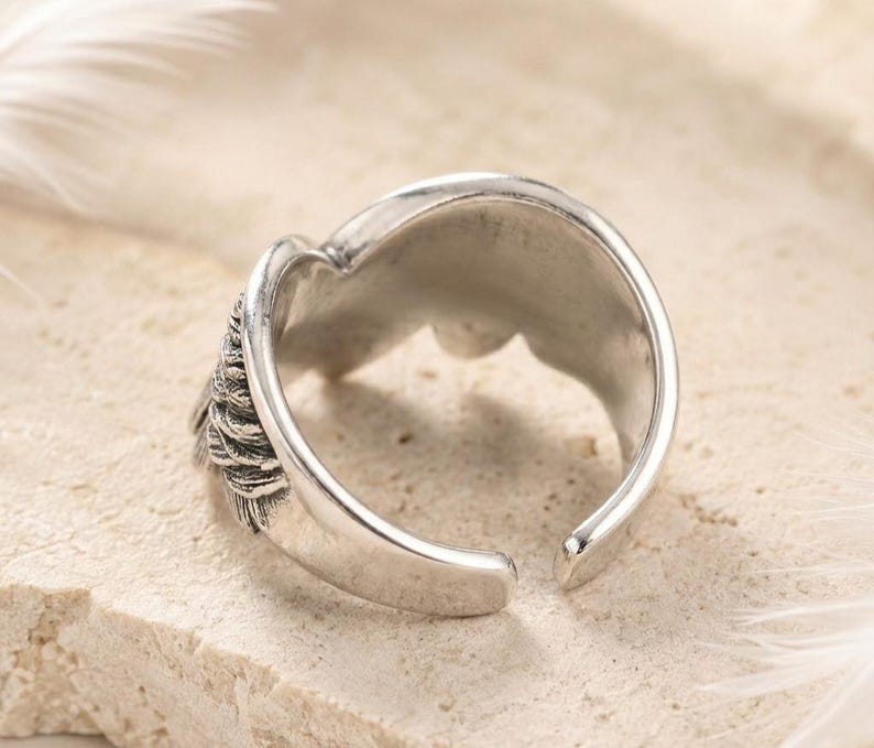 May include: A silver-colored adjustable ring with a detailed feather design on one side. The ring has a smooth, polished finish and an open design, allowing for size adjustment. The ring is set against a neutral background.