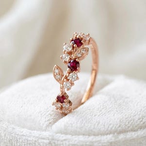 May include: A rose gold ring featuring a vine-inspired design with three round-cut red rubies and multiple small, clear, round-cut white diamonds set along the band. The metal band has a polished finish and a delicate, curved, nature-inspired shape.