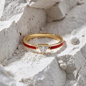 May include: A gold ring with a red enamel band and a triangular, clear gemstone. The ring is set against a textured, white background, highlighting its design and craftsmanship. A modern piece of jewelry.