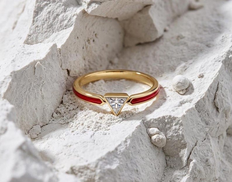 May include: A gold ring with a red enamel band and a triangular, clear gemstone. The ring is set against a textured, white background, highlighting its design and craftsmanship. A modern piece of jewelry.
