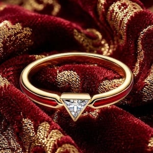 May include: A gold ring with a red enamel band and a triangular, clear gemstone. The ring is set against a rich, red velvet background with gold floral patterns, creating a luxurious aesthetic. The ring's design is modern and elegant.