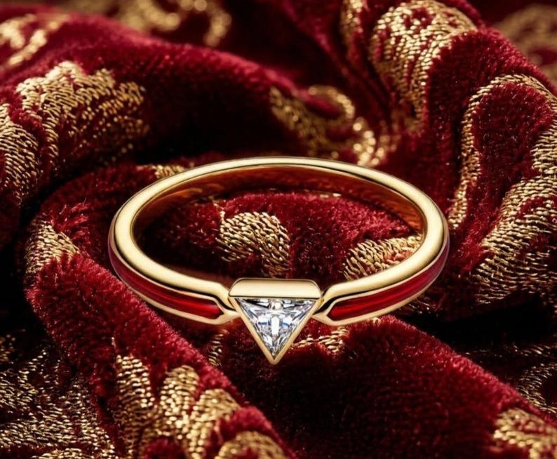 May include: A gold ring with a red enamel band and a triangular, clear gemstone. The ring is set against a rich, red velvet background with gold floral patterns, creating a luxurious aesthetic. The ring's design is modern and elegant.