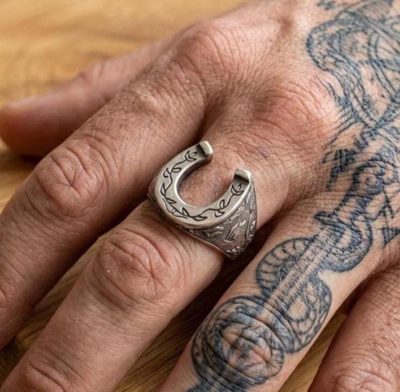 May include: A silver-toned metal ring shaped like a horseshoe, featuring an engraved vine-like pattern along the outer surface. The ring has a polished finish with darkened recessed areas that highlight the intricate floral or leaf-like detailing.