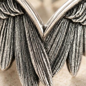 May include: Close-up of a silver-toned metal object shaped like angel wings. The wings have detailed feather textures and a slightly weathered appearance. The design is symmetrical, with each wing mirroring the other.