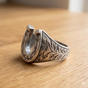 May include: A silver-toned metal ring shaped like a horseshoe. The outer surface of the ring features intricate, engraved leaf and vine patterns that wrap around the sides, with a polished finish and darkened recessed areas to highlight the details.