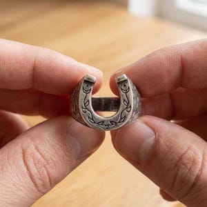 May include: A silver-toned metal ring shaped like a horseshoe. The surface of the ring features engraved floral or vine-like patterns along the outer edges and the curved top section, with a dark, oxidized finish within the recessed engravings.