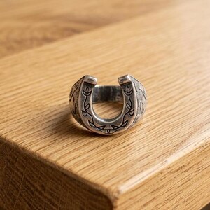 May include: A silver-toned metal ring shaped like a horseshoe, featuring an engraved vine and leaf pattern along the front surface. The band has a polished finish with darkened recessed areas that highlight the intricate botanical design details.