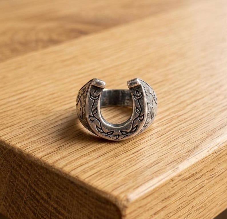 May include: A silver-toned metal ring shaped like a horseshoe, featuring an engraved vine and leaf pattern along the front surface. The band has a polished finish with darkened recessed areas that highlight the intricate botanical design details.