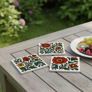Könnte beinhalten: Drei quadratische Keramikuntersetzer mit floralem Design. Jeder Untersetzer hat einen weißen Hintergrund mit roten, gelben und blauen Blumen und grünen Blättern. Die Untersetzer sind auf einem Holztisch angeordnet.