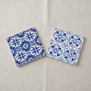 May include: Two square ceramic coasters with a blue and white floral pattern. The design features stylized flowers and leaves in shades of blue against a white background. The coasters are placed on a neutral-colored surface.