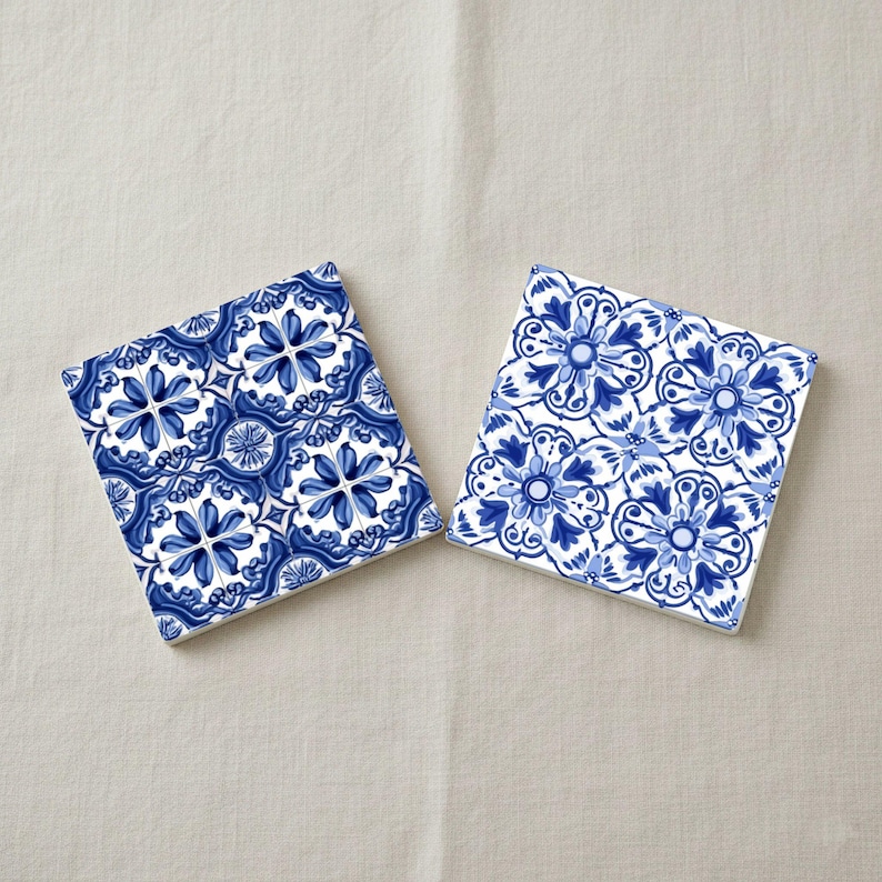 May include: Two square ceramic coasters with a blue and white floral pattern. The design features stylized flowers and leaves in shades of blue against a white background. The coasters are placed on a neutral-colored surface.