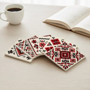 Peut inclure: Ensemble de dessous de verre carrés blancs ornés de motifs de broderie au point de croix rouges et noirs. Une tasse à café blanche et un livre ouvert sont visibles. Les dessous de verre sont disposés en éventail.