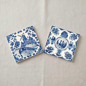 May include: Two square ceramic coasters with blue and white Dutch windmill and floral designs. One coaster features a windmill over a bridge and river. The other coaster has a circular ship design surrounded by floral patterns.