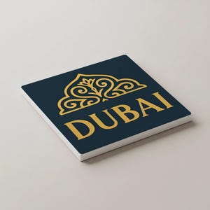 May include: A square coaster with a navy blue surface and a gold design featuring the word "DUBAI". The design includes an ornate, stylized emblem above the word. The coaster has a white border.