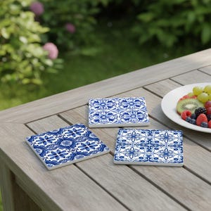 May include: Three square ceramic coasters with a blue and white floral pattern are arranged on a wooden table. The coasters have a glossy finish and are placed near a plate of fresh fruit. The background shows a green lawn and foliage.