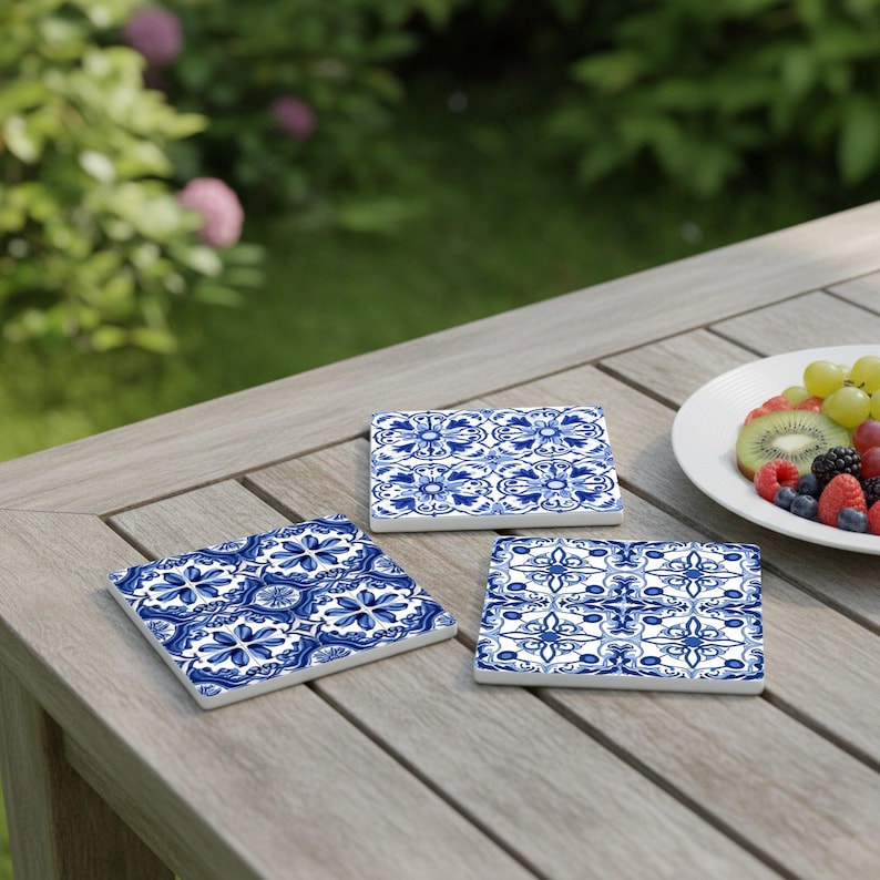 May include: Three square ceramic coasters with a blue and white floral pattern are arranged on a wooden table. The coasters have a glossy finish and are placed near a plate of fresh fruit. The background shows a green lawn and foliage.