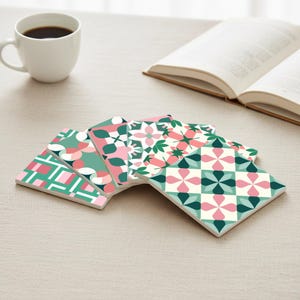 Puede incluir: Un juego de seis posavasos cuadrados de cerámica con varios patrones geométricos y florales en tonos rosa, verde y blanco. Una taza de café blanca y un libro abierto están en el fondo.