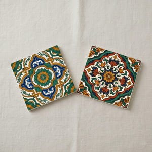 May include: Two square ceramic coasters with detailed floral designs. One coaster displays a green, blue, and yellow pattern, the other a red, green, and yellow design. Both are set against a white backdrop.