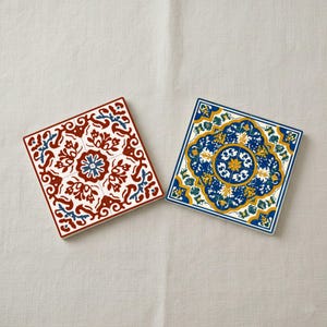 May include: Two square ceramic coasters with detailed floral patterns. One has a red, white, and blue design, the other a blue, yellow, and white design. The coasters are displayed on a neutral-coloured surface.
