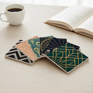 May include: Six square coasters with diverse geometric and art deco patterns. The coasters display black, white, pink, teal, navy, and gold hues. A white coffee mug and an open book are also visible in the image.