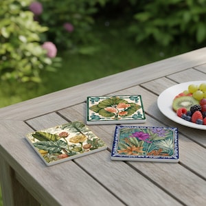Puede incluir: Tres posavasos cuadrados de cerámica con diseños botánicos sobre una mesa de madera. Cada posavasos presenta una ilustración diferente de plantas tropicales con colores vibrantes y bordes decorativos. Un plato de fruta está cerca.