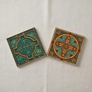May include: Two square coasters with detailed patterns. One coaster has the text "SAN FRANCISCO" in teal, set within a teal and gold geometric design. The other coaster features a circular design with teal and gold detailing.