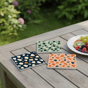 Könnte beinhalten: Drei quadratische Untersetzer mit Terrazzo-Mustern in Marineblau, Pfirsich und Grün liegen auf einem Holztisch. Die Untersetzer haben einen weißen Rand und ein farbenfrohes Design aus unregelmäßigen Formen. Ein Teller mit Obst befindet sich im Hintergrund.