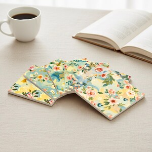 Könnte beinhalten: Ein Set aus sechs quadratischen Keramikuntersetzern mit floralem Design. Die Untersetzer haben eine Pastellfarbpalette mit gelben, blauen und rosa Blumen. Eine weiße Kaffeetasse und ein offenes Buch befinden sich im Hintergrund.